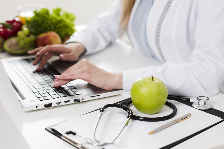 braços e mãos de uma mulher branca digitando em um notebook, em cima de um local branco, com verduras do seu lado direito e uma maça verde em cima de prancheta com papeis e um estetoscópio em volta