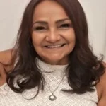 Mulher morena de cabelos médios lisos e blusa branca em um fundo branco.