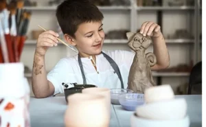 Menino sorridente sentado em uma mesa de artesanato, moldando uma escultura de gato com argila. Ele segura um pincel em uma mão e a escultura na outra. Ao fundo, há prateleiras com materiais de arte e cerâmica. A criança está se divertindo durante uma atividade artística.