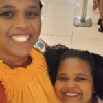 Uma selfie alegre de uma mulher negra e com cabelo trançado com uma blusa laranja e uma menina negra sorridente de cabelos cacheados. Ao fundo, um ambiente movimentado e desfocado.