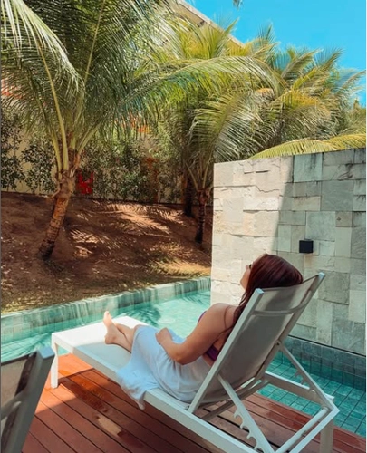 Mulher de pele morena e cabelo liso castanho, deitada em espreguiçadeira à beira de uma piscina, rodeada de palmeiras, apreciando o sol.