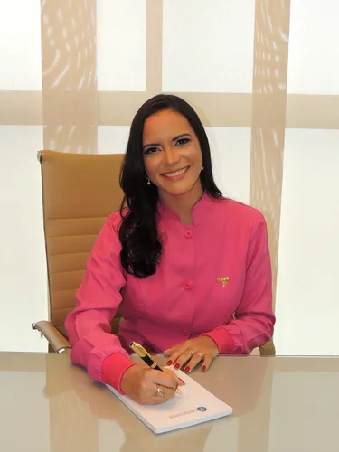 Odontopediatra Andrea Chagas vestido de jaleco rosa e sentada numa cadeira bege assinando um papel