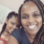 Mulher e criança negras sorrindo para uma selfie, ambas com cabelos trançados, sentadas lado a lado.