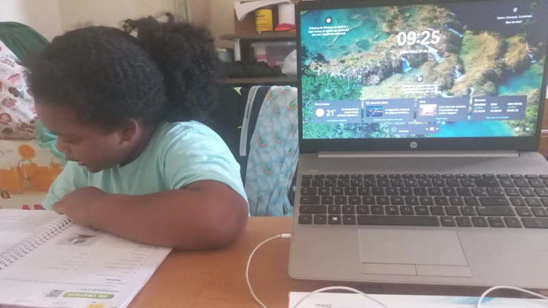 Criança escrevendo em um caderno sobre uma mesa com um notebook ao lado, exibindo tela ligada com horário e widgets.