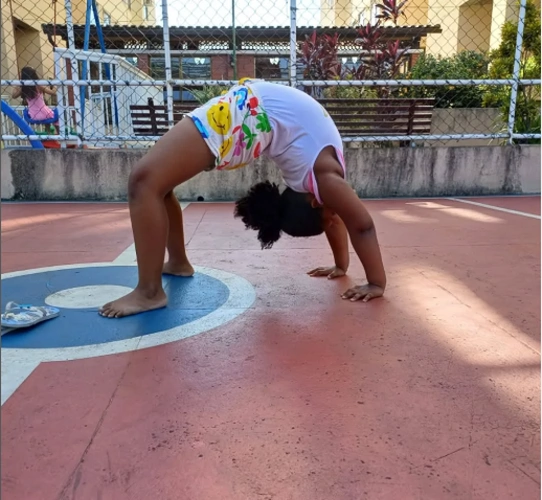 Uma menina negra descalça fazendo uma ponte de ginástica em uma quadra poliesportiva. Ela está de costas, com os braços e pernas estendidos, mostrando sua flexibilidade.