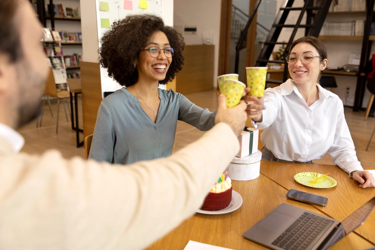 Três pessoas brindam com copos descartáveis em um ambiente de escritório, sorrindo ao redor de uma mesa com bolo e presentes.