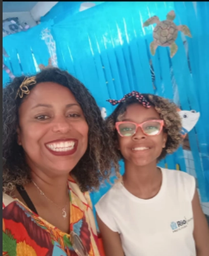 Mulher negra com cabelos cacheados e pintados de loiro nas pontas e blusa colorida ao lado de uma menina negra com uma blusa branca e óculos