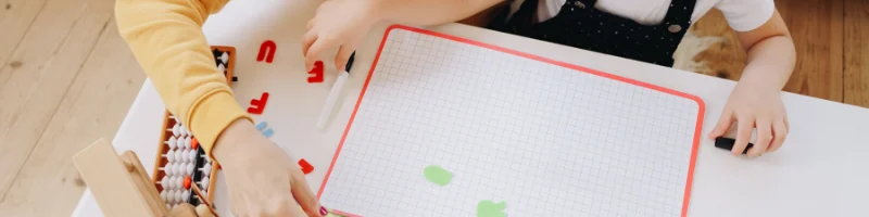 Adulto e criança brincando em uma mesa com quadro magnético de letras coloridas, ábaco de madeira e papéis coloridos, em ambiente educativo e lúdico para aprendizado infantil