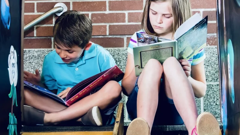 "Duas crianças sentadas em uma escada lendo livros, representando um momento tranquilo e focado de homeschooling."
