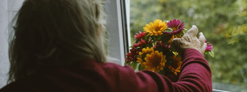 Pessoa idosa de cabelos grisalhos, vestindo um suéter vermelho, coloca um vaso de flores coloridas no parapeito da janela, olhando para fora. A imagem transmite um momento de contemplação e calma. Acessibilidade visual: a cena enfatiza a importância de ambientes iluminados e acessíveis para pessoas idosas