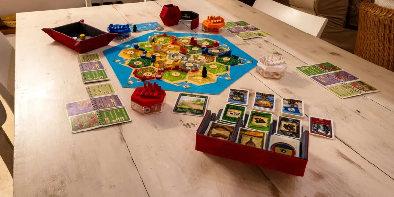 Jogo de tabuleiro à mesa, esta tem a cor clara e o jogo de cartas é colorido com um tabuleiro azul com peças pequenas sob o mesmo.