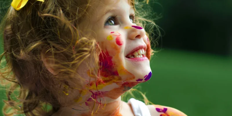 arte 2 Criança sorridente com cabelos cacheados e laço amarelo, com o rosto, braços e roupa cobertos por tinta colorida, em um ambiente ao ar livre.